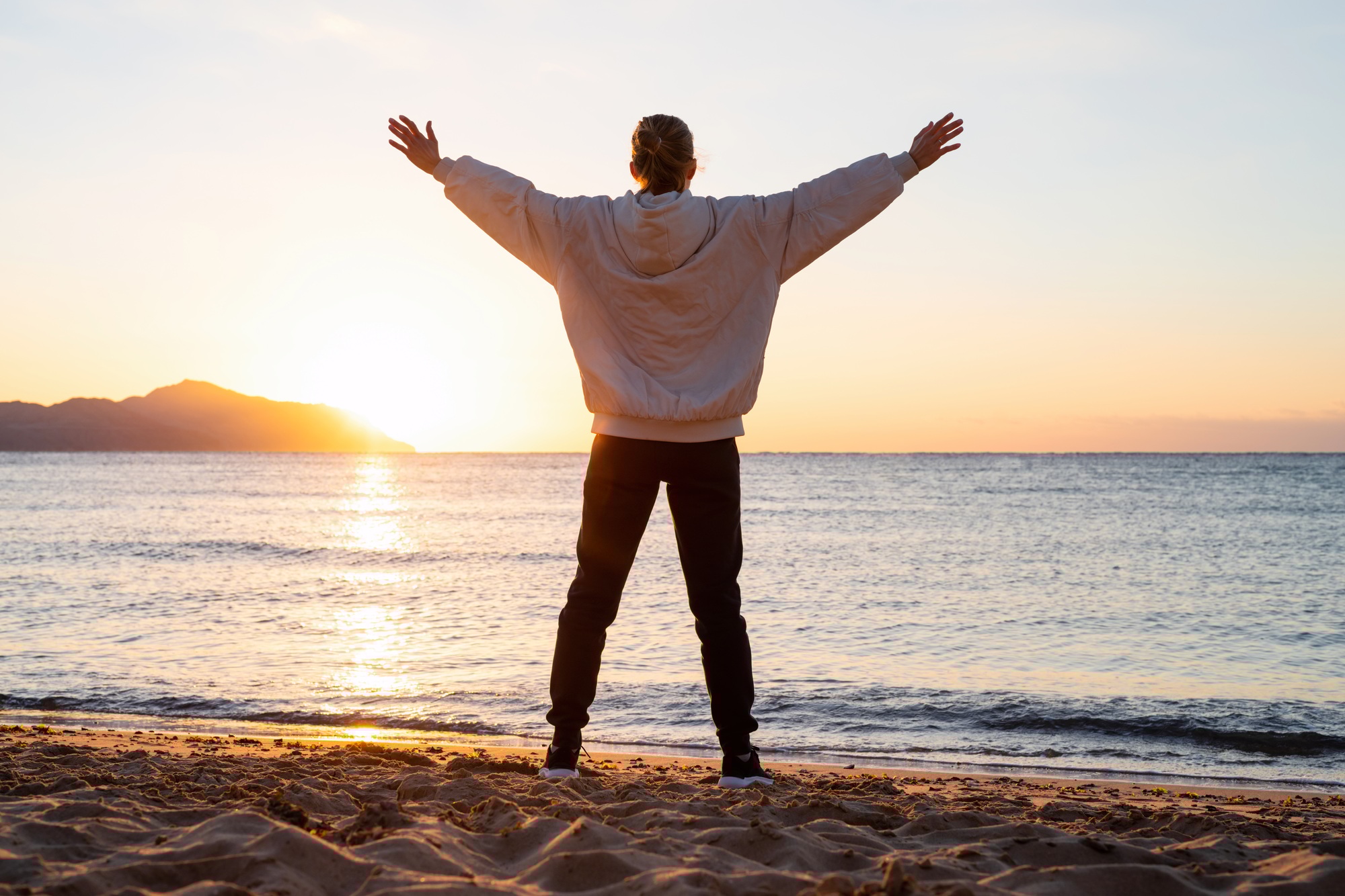 Woman working out at the beach at sunrise, looking at the horizon with arms outstretched